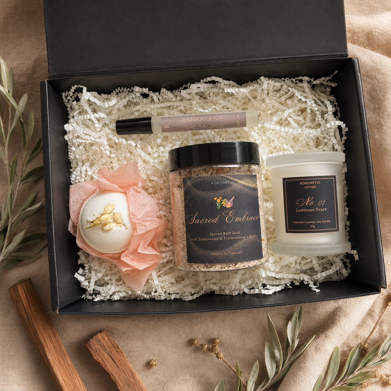 An open black gift box filled with white shredded paper and a curated spa gift set, including a jar labeled Sacred Embrace bath soak, a candle labeled No. 07 Luminous Grace a mini space mist, and a round bath bomb wrapped in peach tissue paper.