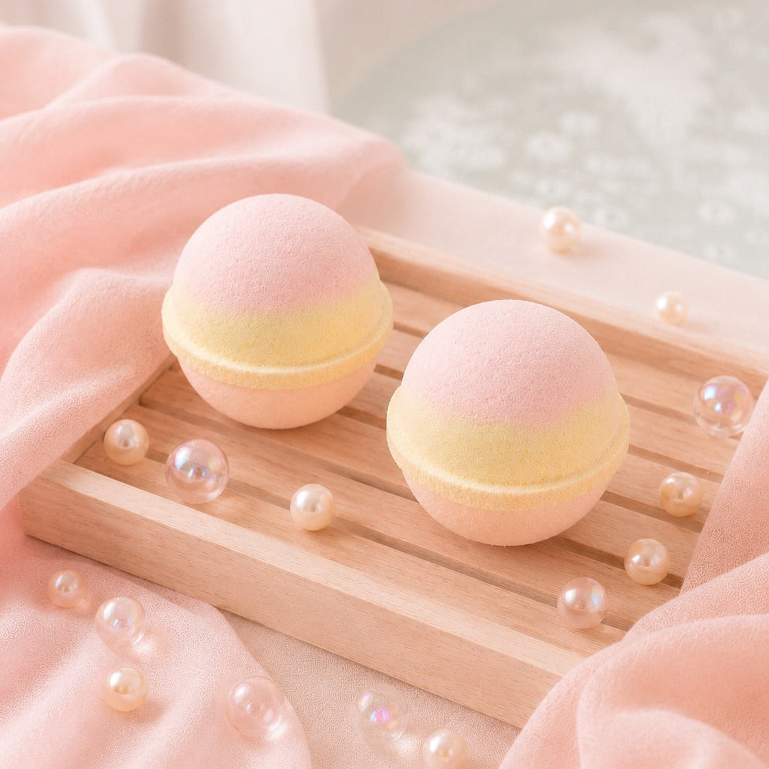 Two pink bath bombs on a wooden tray with pearls, surrounded by soft pink and white background.