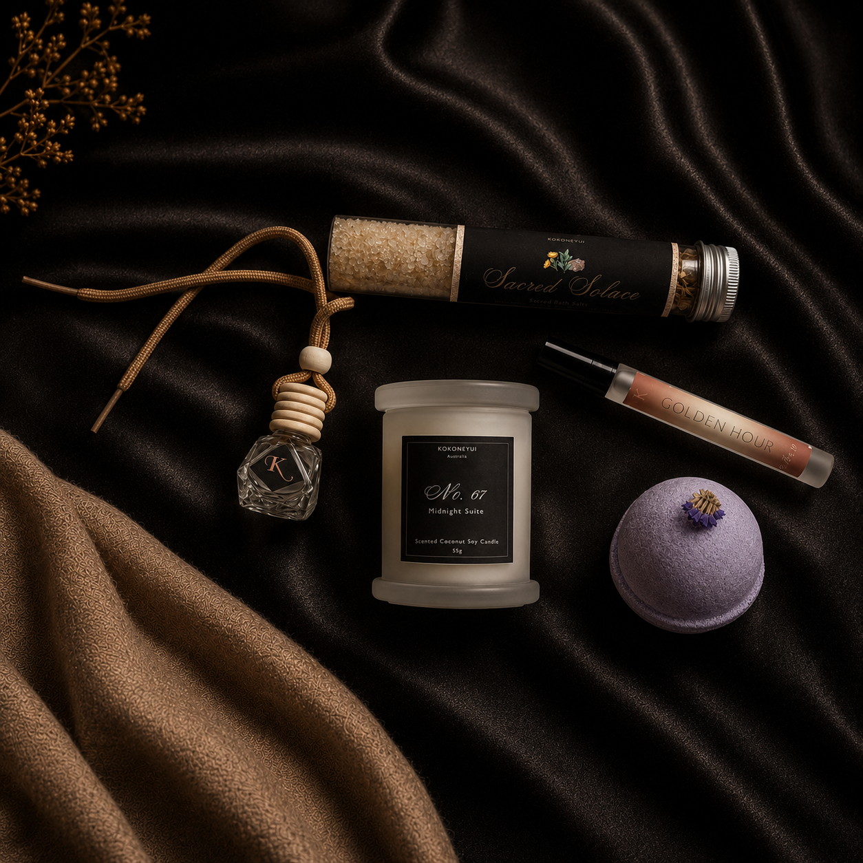A styled flat-lay of multiple premium self-care and home fragrance products on dark draped satin fabric: a jar candle labeled &