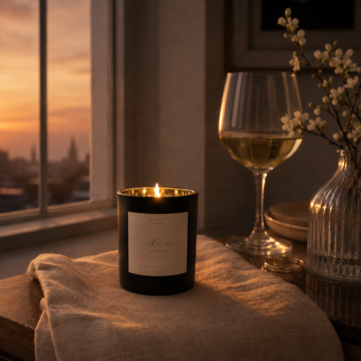 Candle with a label, wine glass, and vase on a table by a window at sunset.