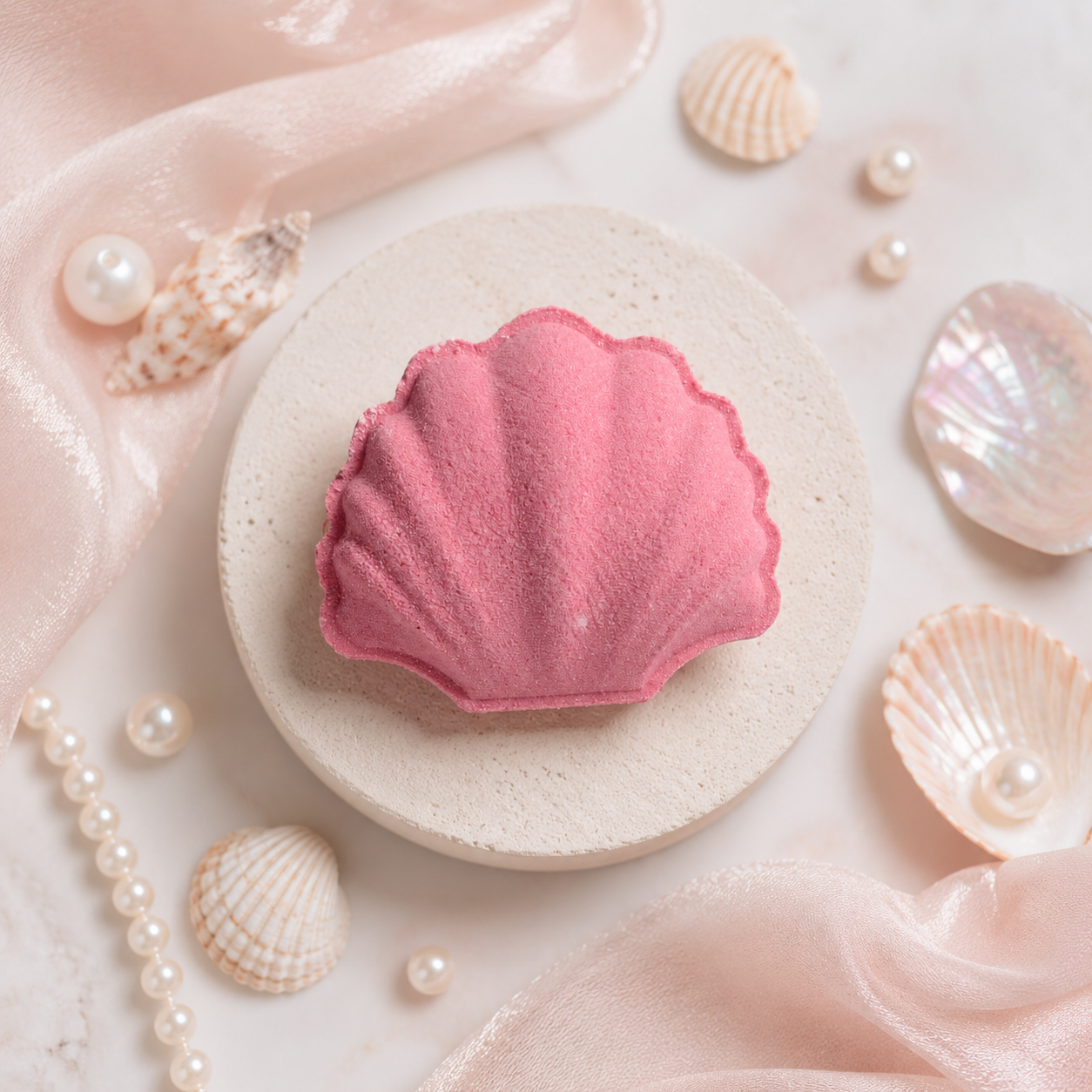 Pink shell-shaped bath bomb on a white plate with seashells and pearls around it.