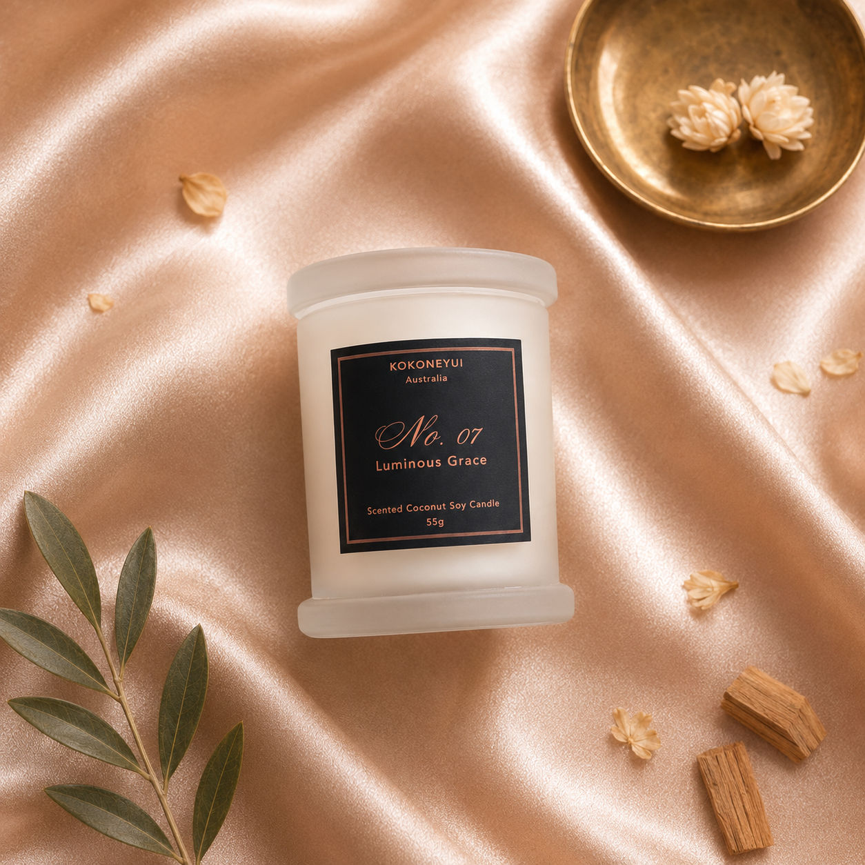 A frosted glass jar candle labeled 'KOKONEYUI No. 07 Luminous Grace' sits on draped champagne satin with scattered dried flowers, a green leaf sprig, and small wooden pieces.