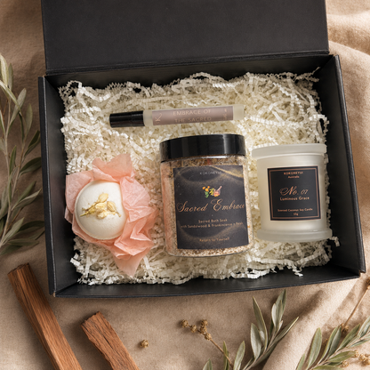 An open black gift box filled with white shredded paper and a curated spa gift set, including a jar labeled Sacred Embrace bath soak, a candle labeled No. 07 Luminous Grace a mini space mist, and a round bath bomb wrapped in peach tissue paper.