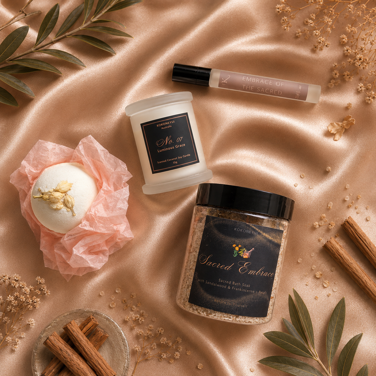 A styled flat-lay of spa and self-care products on champagne satin fabric, including a jar labeled Sacred Embrace bath soak, a frosted candle, a mini space mist, and a white bath bomb wrapped in tissue paper, surrounded by cinnamon sticks, dried flowers, and green leaves.