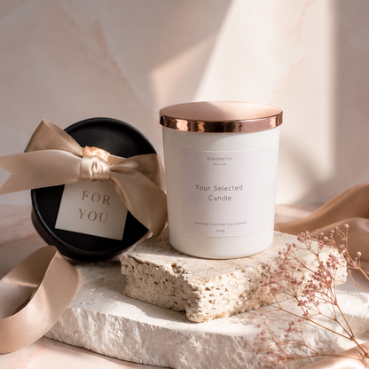 White candle with rose gold lid on a stone surface, accompanied by a black gift box with a bow.