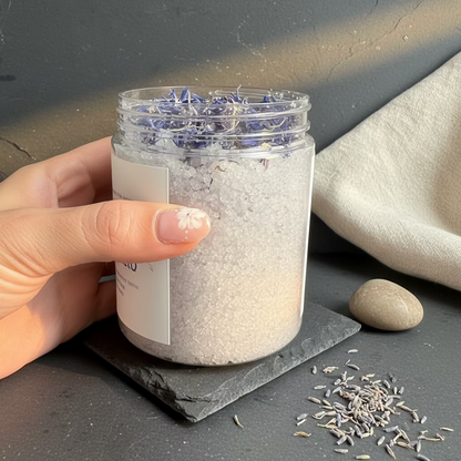 Hand holding a jar of Scrub with blue Cornflower petals on a dark surface