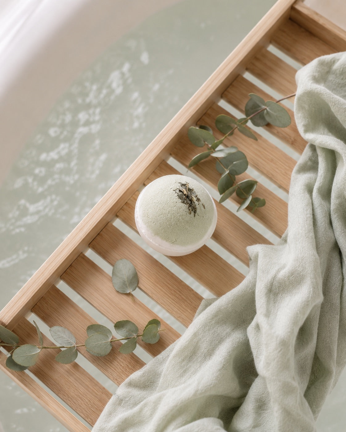 Wooden tray with a bath bomb, eucalyptus leaves, and a towel over a bathtub.