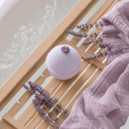 Lavender bath bomb on a wooden tray with lavender flowers and a towel in a bathtub.