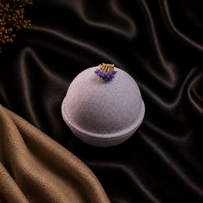 A round lavender bath bomb with a small dried flower, resting on draped black satin fabric with a brown textured cloth in the corner.