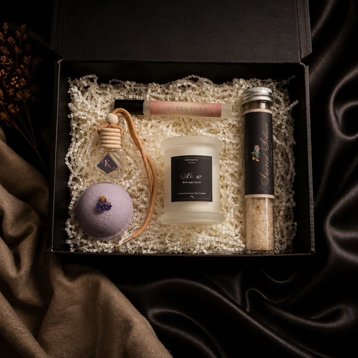 An open black gift box filled with white crinkle paper and spa-style items, including a mini candle, a bath salt tube, a bath bomb, a car diffuser, and a space mist, arranged on dark satin fabric.