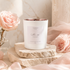 White candle with rose gold interir on a stone surface surrounded by pink roses