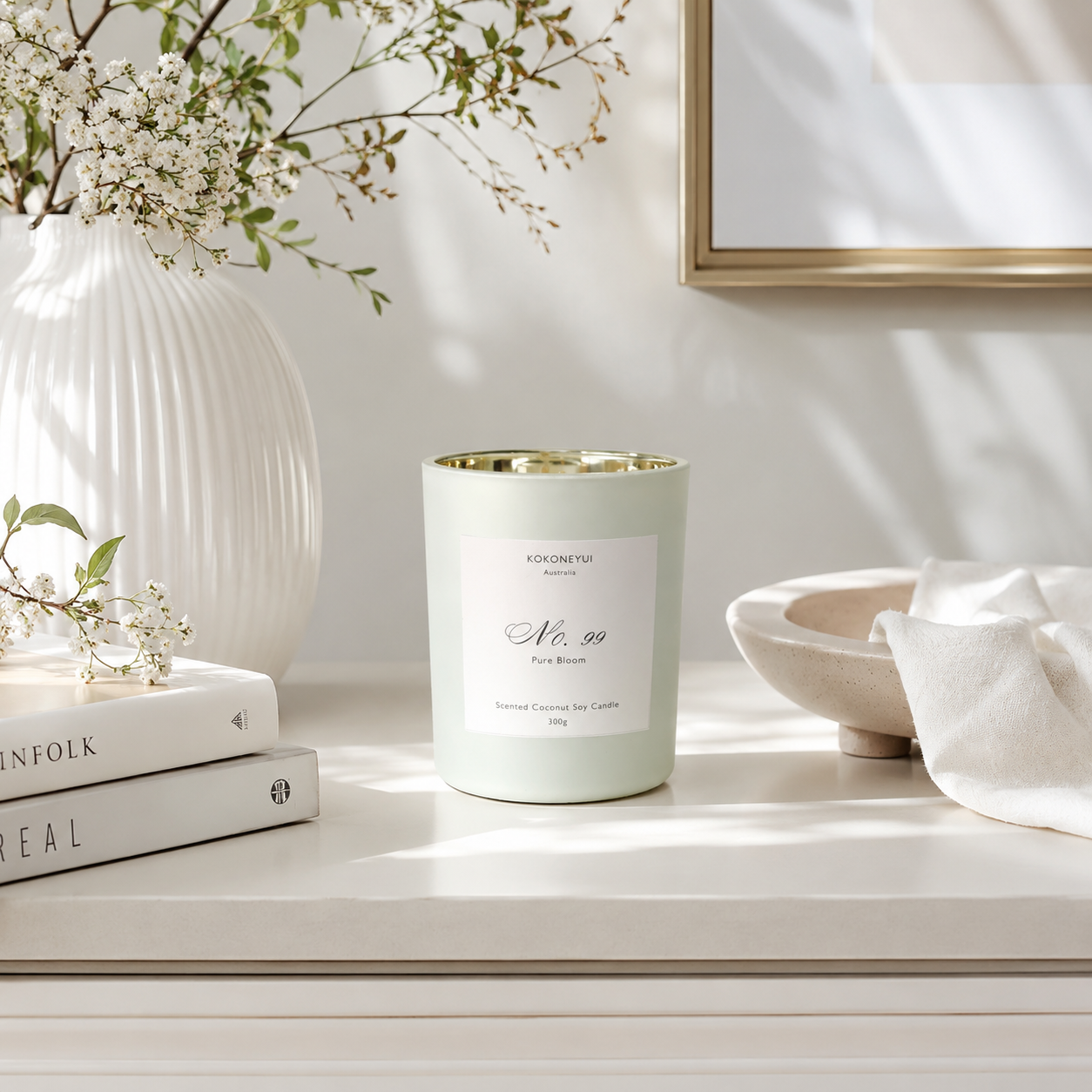 Candle with a white label on a surface with books, a vase, and flowers in the background
