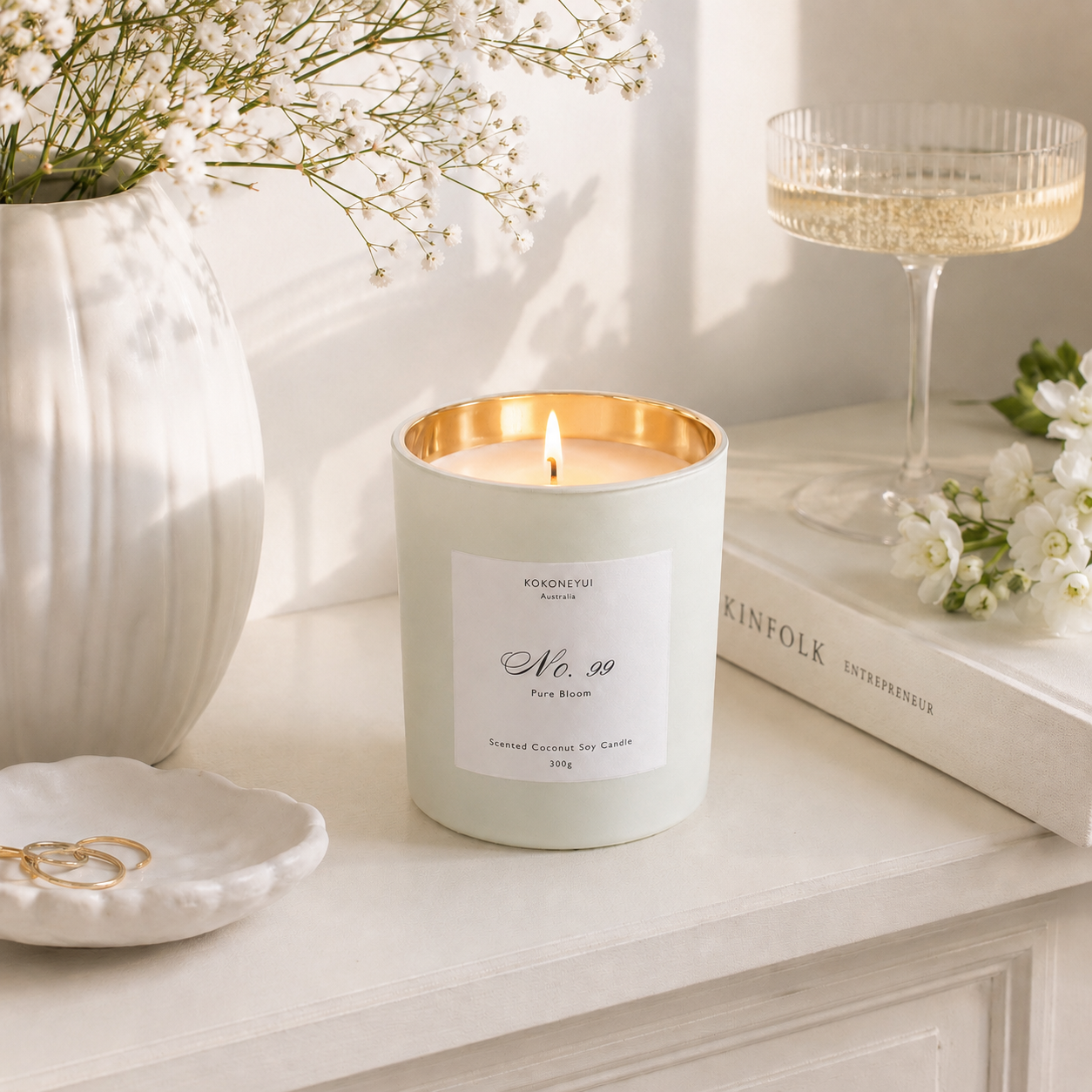 Lit candle in a decorative container on a white surface with flowers and books in the background