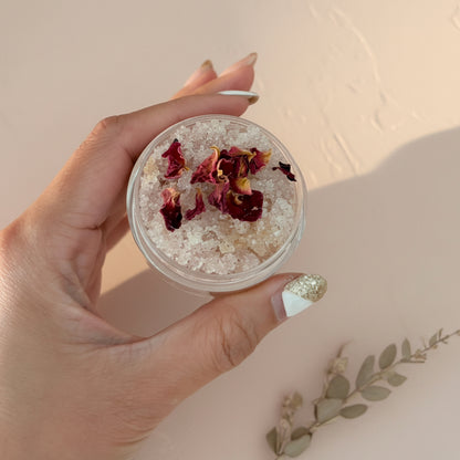 Hand holding a scrub container with dried flowers against a plain background