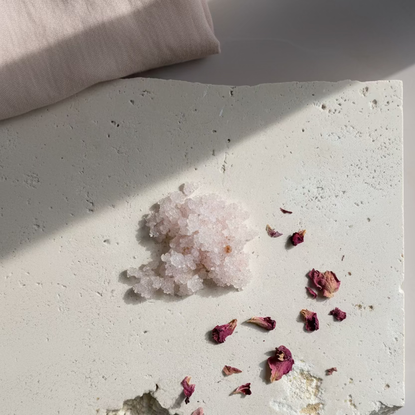 Pink Body Scrub and dried rose petal arranged on a textured white stone surface for a soft botanical self-care ritual. 