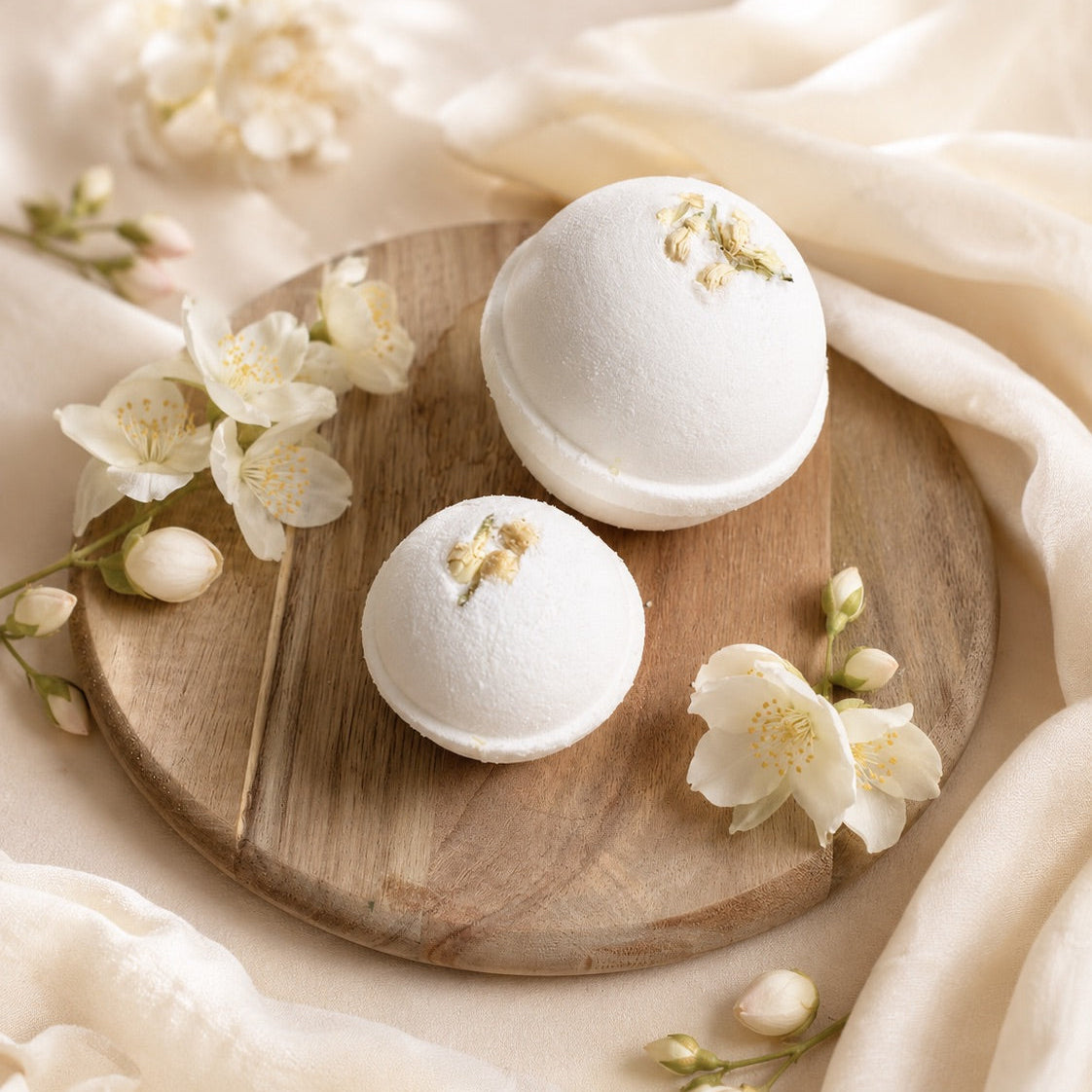 Two white bath bombs with gold accents on a wooden board surrounded by white flowers.