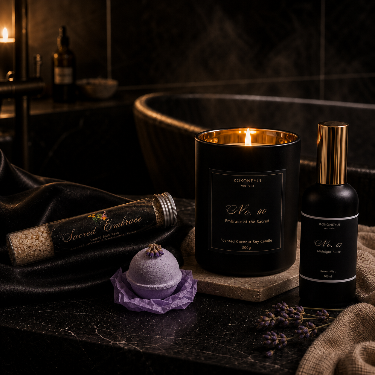 A styled product arrangement on a dark marble surface featuring a black candle with a gold-toned inner rim and lit flame, a black room mist bottle with a gold cap, a bath soak tube labeled Sacred Embrace, a lavender x Ylan Ylan bath bomb on purple tissue paper, and draped black satin fabric. 