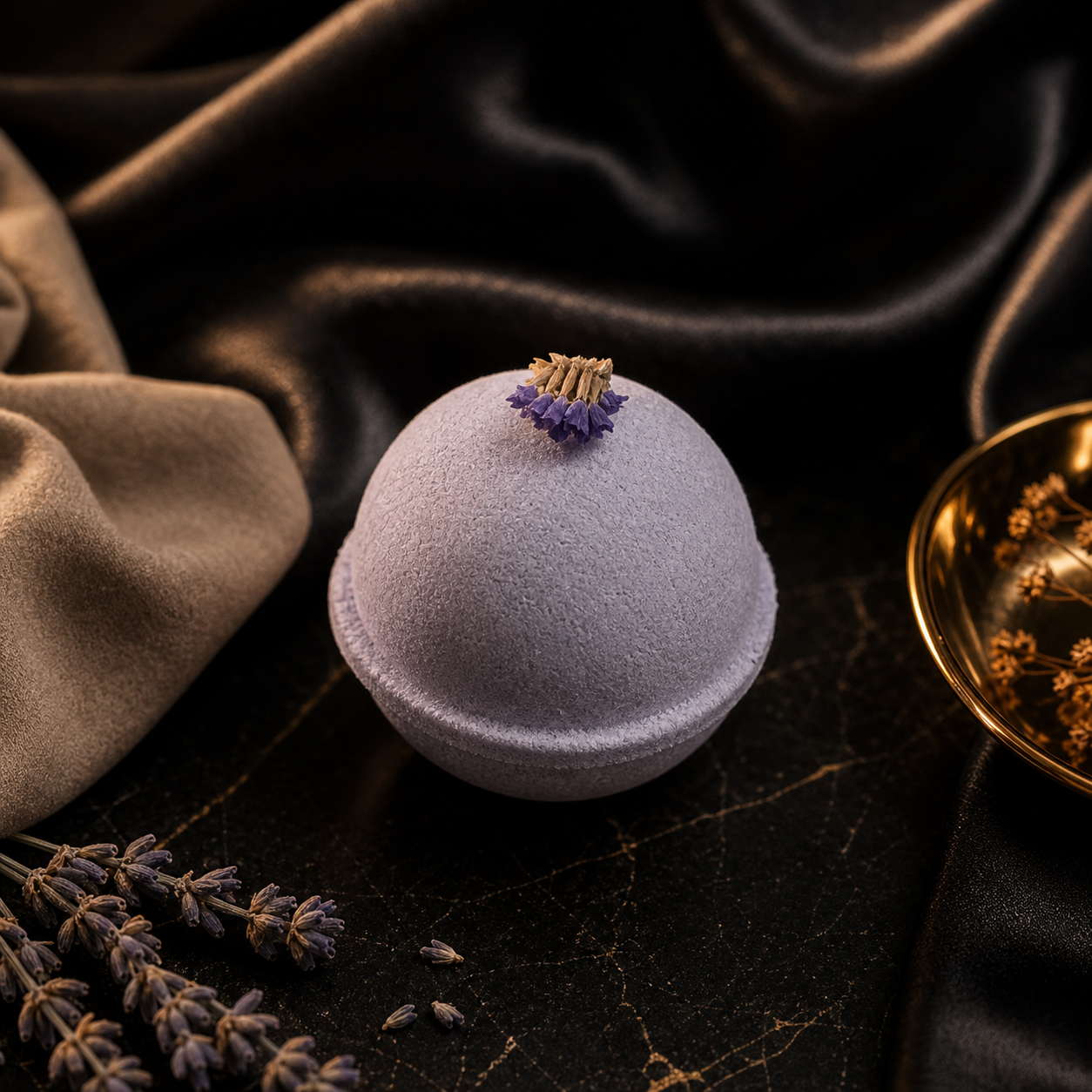 A lavender bath bomb with a small purple flower, resting on a dark draped satin surface with fabric and dried lavender props.